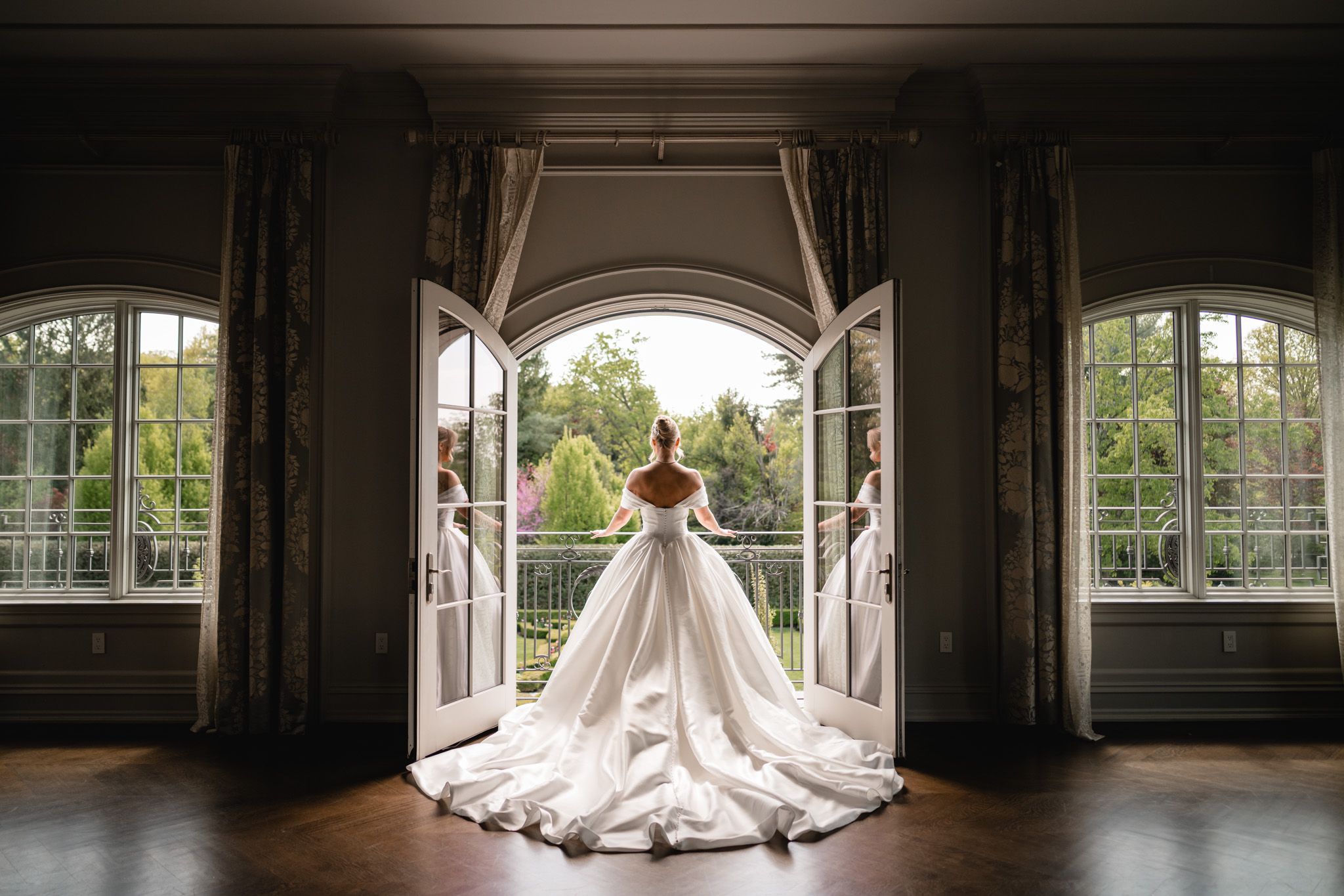 Bride in elegant white ball gown stands in arched doorway overlooking lush garden at luxury wedding venue.