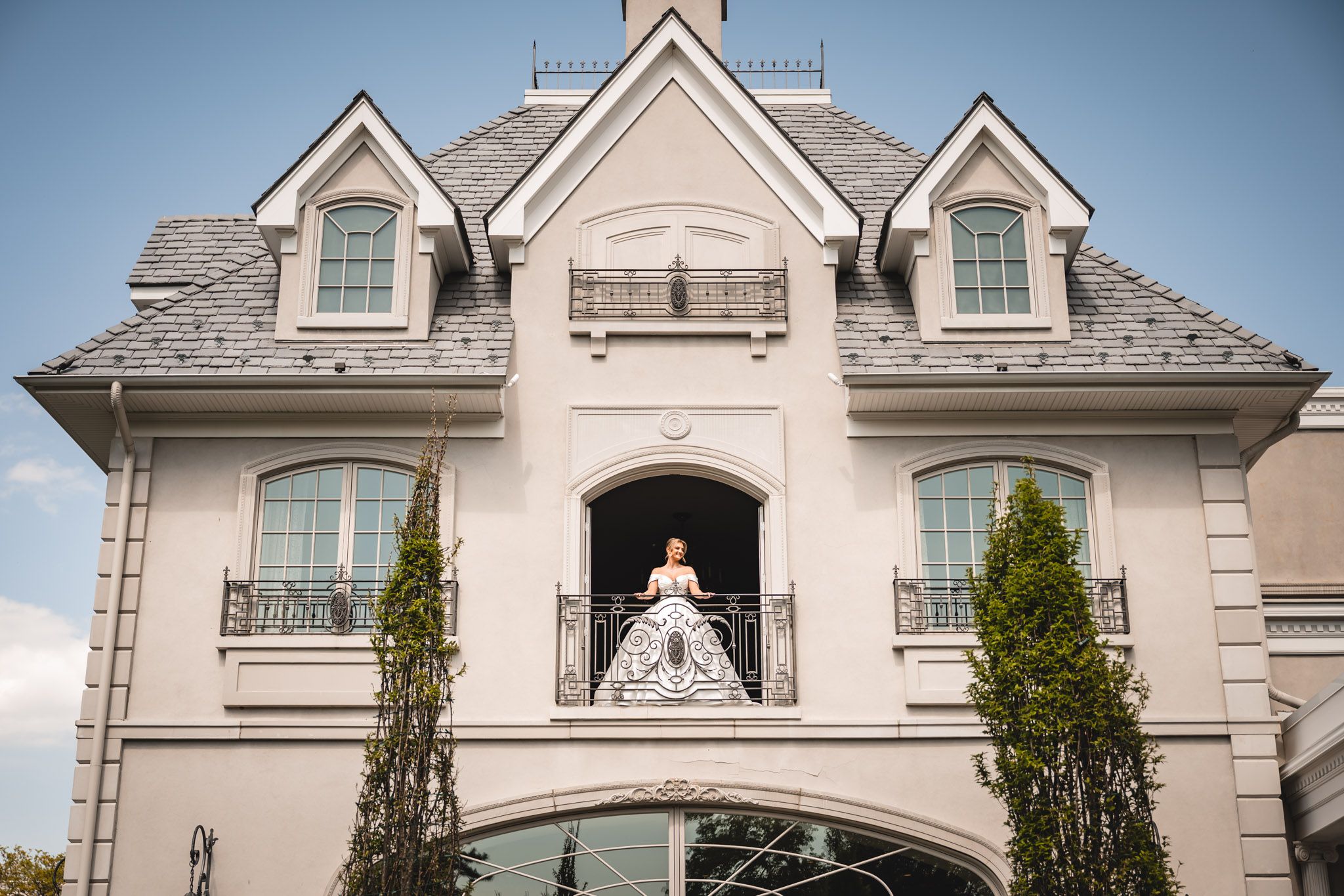 Bride in white gown standing on ornate balcony of elegant French chateau-style mansion with gray roof.