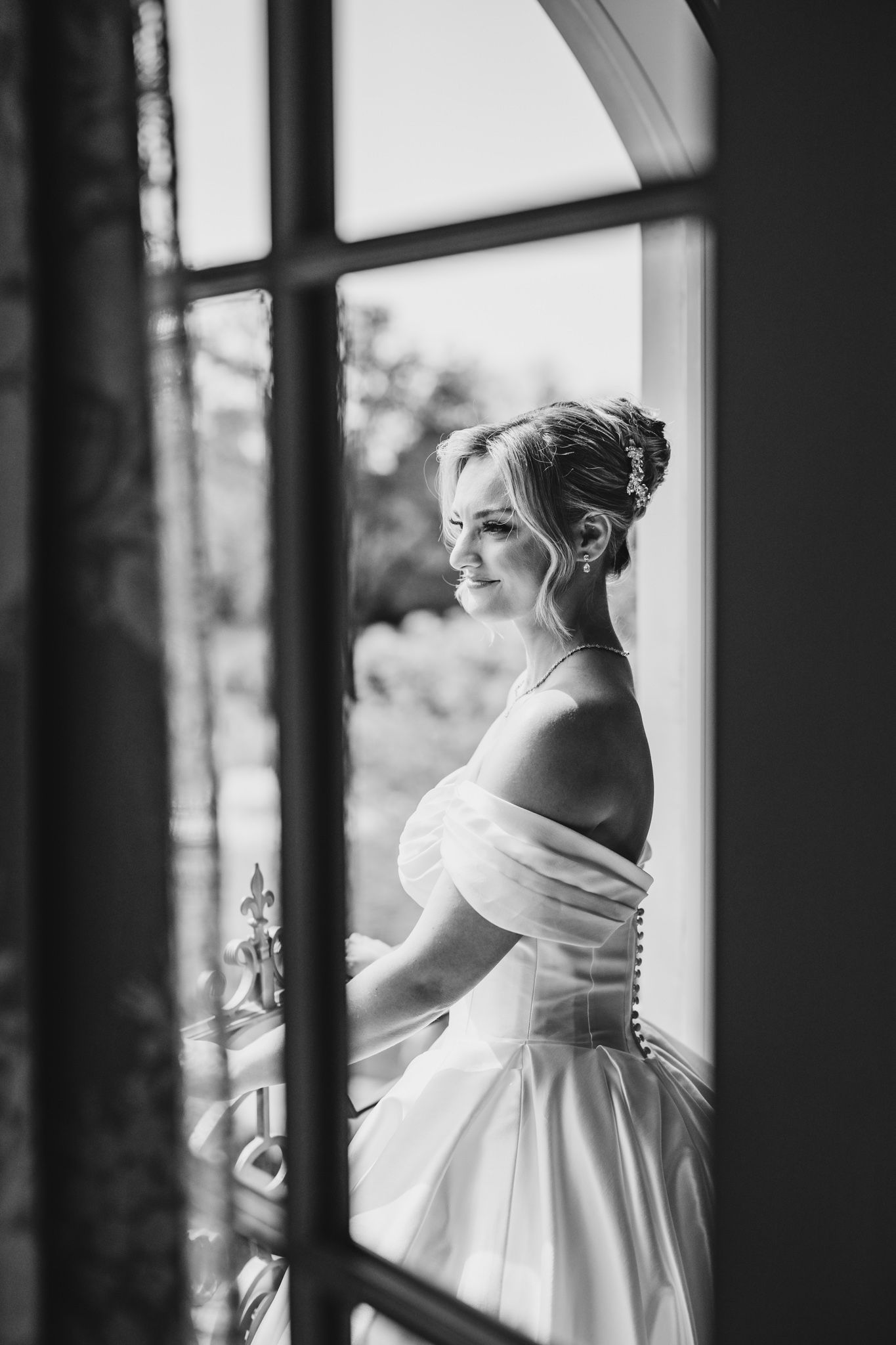 Bride in off-shoulder white gown smiling softly by a window in a black and white wedding portrait.