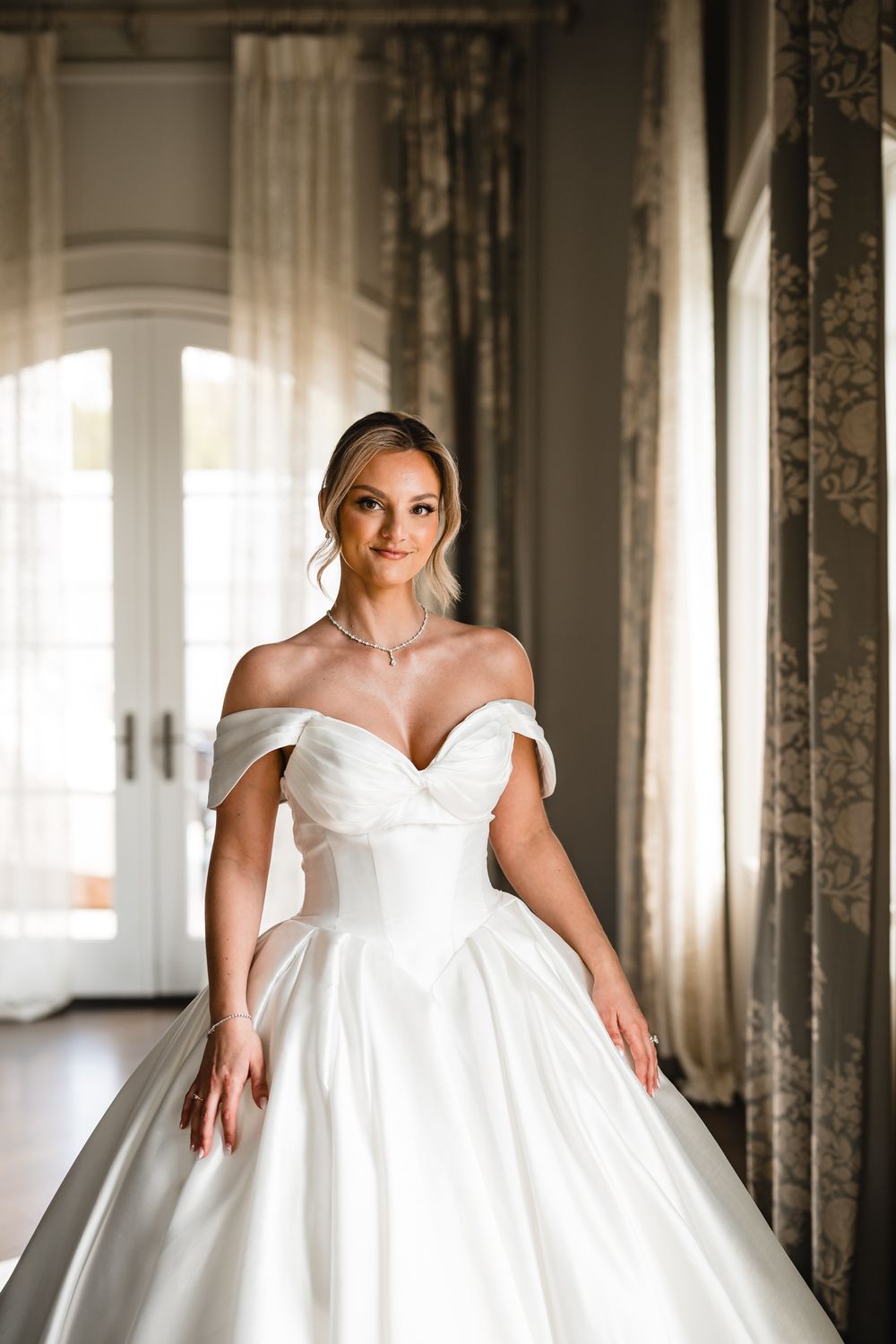 Elegant bride in off-shoulder white ballgown with sweetheart neckline and diamond necklace posing indoors.