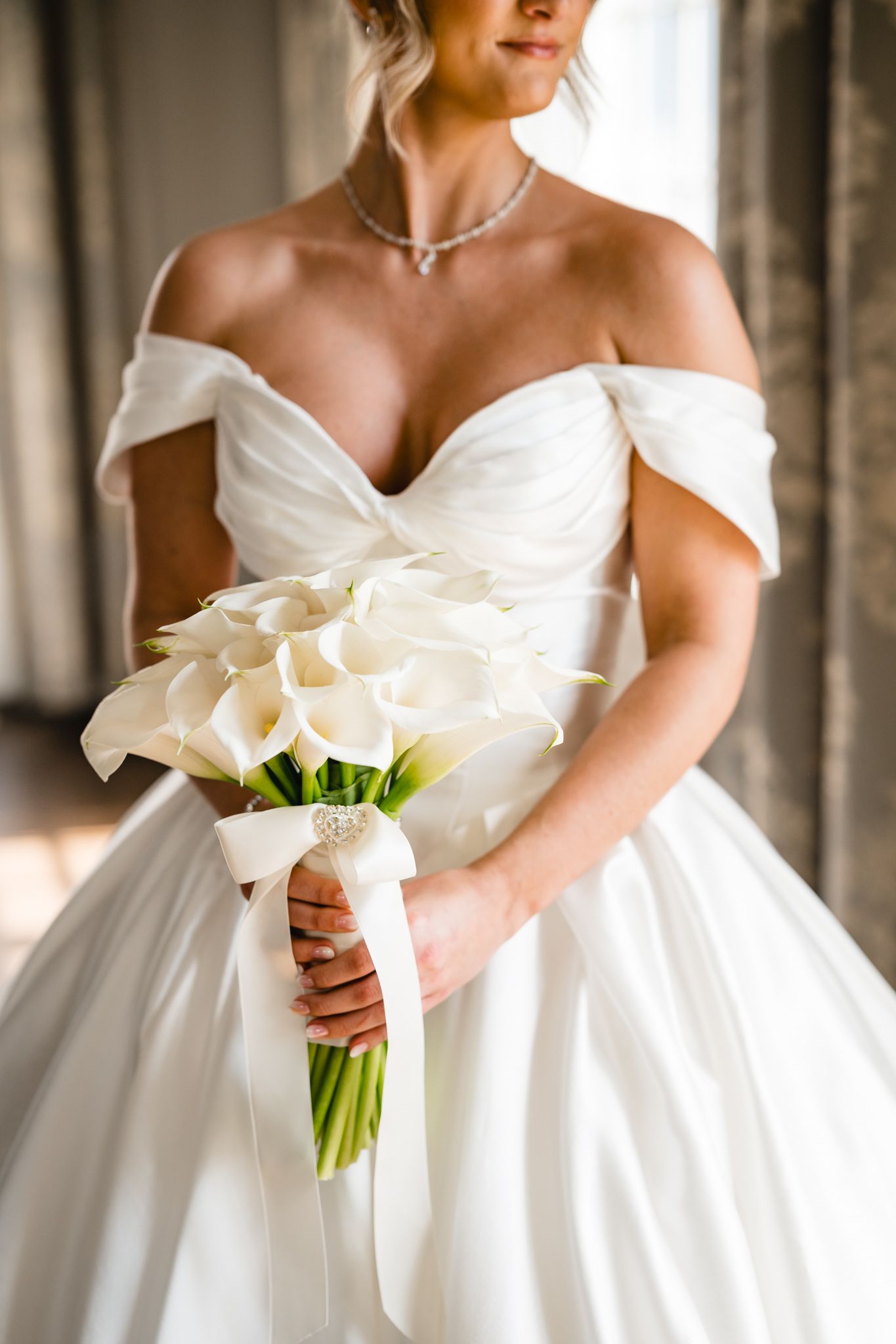 Bride holding white calla lily bouquet with ribbon in off-shoulder satin ballgown and diamond necklace.