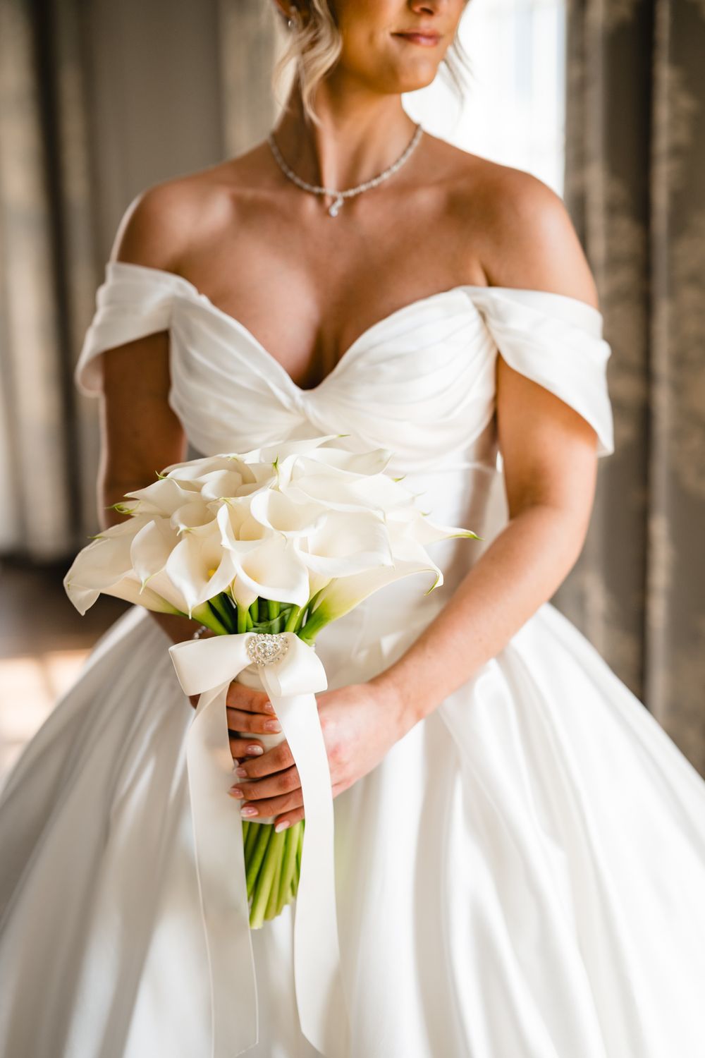 Bride holding white calla lily bouquet with ribbon in off-shoulder satin ballgown and diamond necklace.
