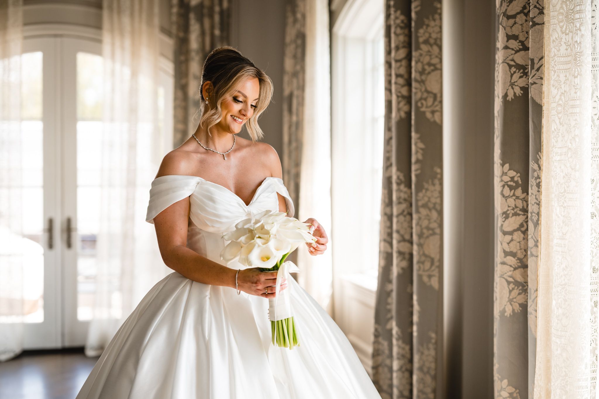 Smiling bride in off-shoulder white ball gown holds white calla lily bouquet by sunlit window.