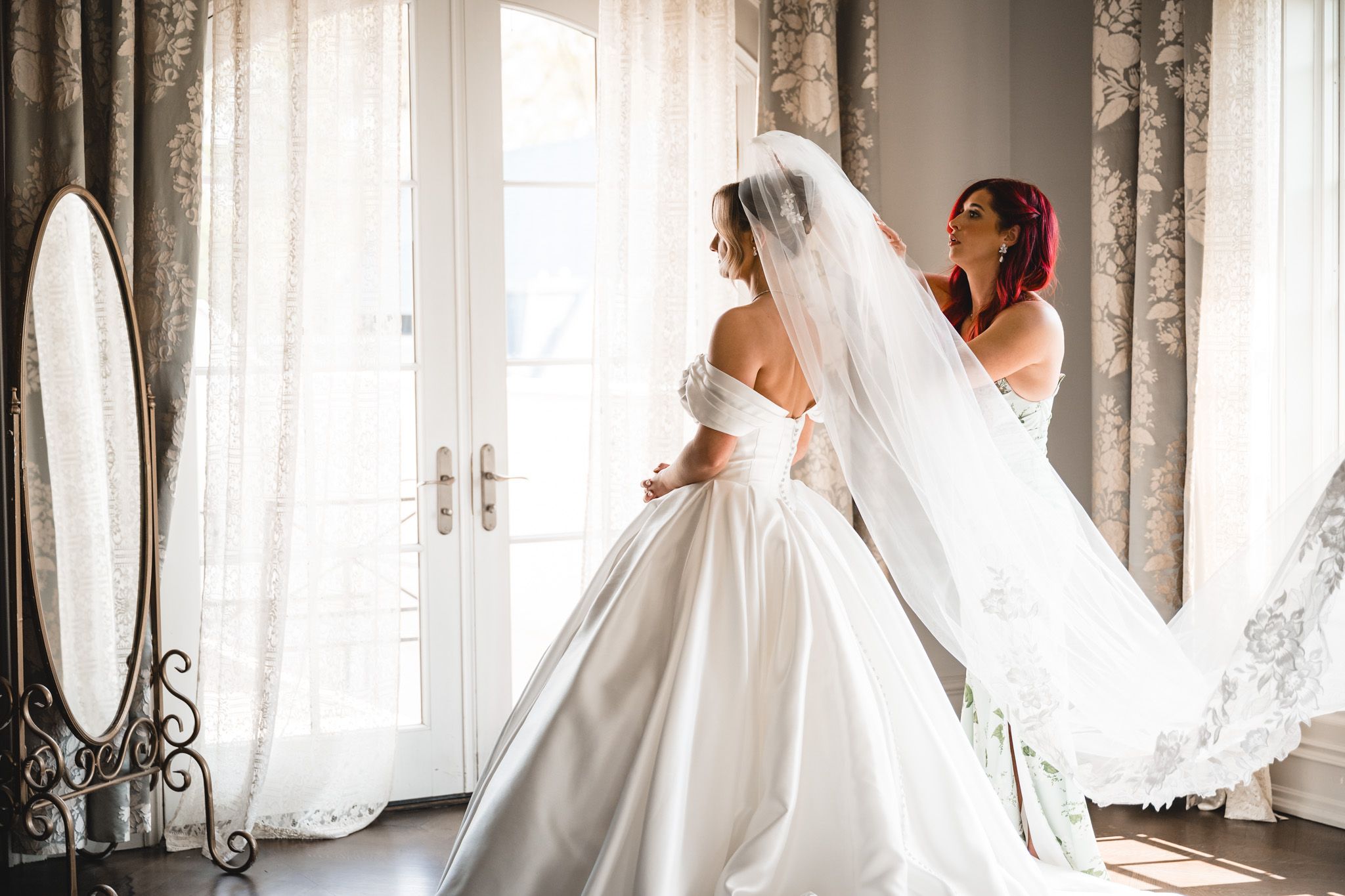 Bridesmaid helping bride arrange long flowing veil near bright window before wedding ceremony.