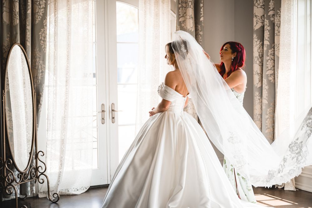 Bridesmaid helping bride arrange long flowing veil near bright window before wedding ceremony.