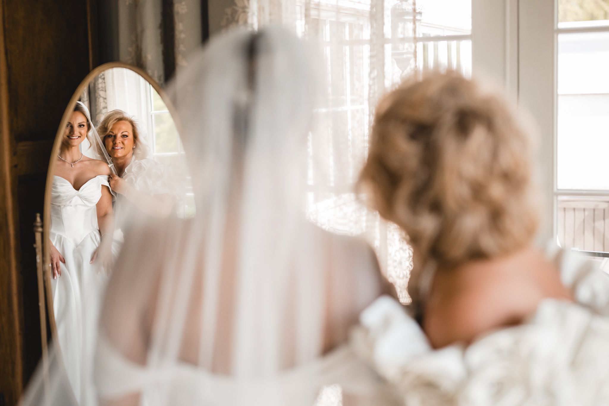 Bride in white gown views her reflection in an oval mirror while bridesmaid stands behind her.