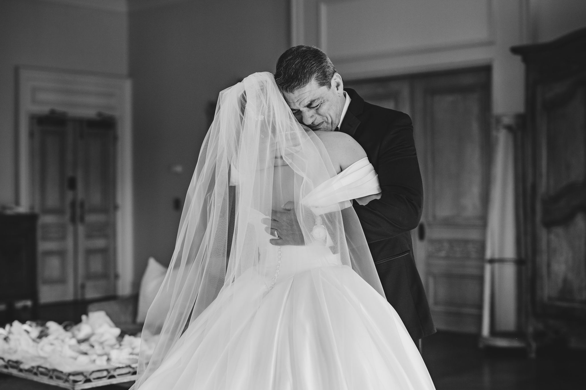 Black and white photo of groom tenderly embracing bride in wedding gown and veil indoors.
