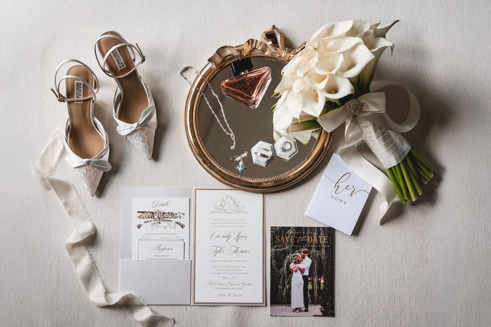 Wedding flat lay featuring white heels, calla lily bouquet, invitations, jewelry, and accessories on neutral background.