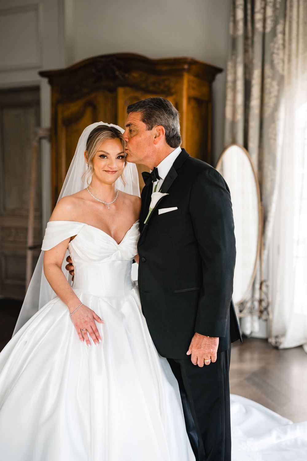 A bride in a white ball gown and veil receives a forehead kiss from a man in a black tuxedo in an elegant room.