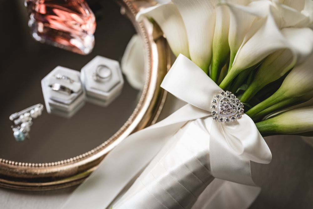 Elegant wedding bouquet with white calla lilies, silver heart brooch, and ribbon beside jewelry boxes and rings on a tray.