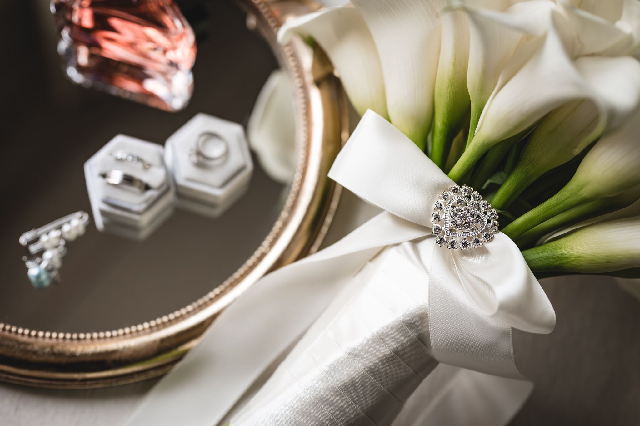 Elegant wedding bouquet with white calla lilies, silver heart brooch, and ribbon beside jewelry boxes and rings on a tray.