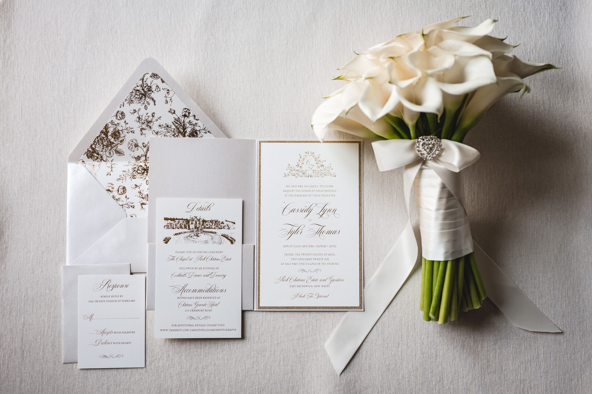 Elegant wedding invitation suite with gold details, envelope liner, and white calla lily bouquet on gray background.