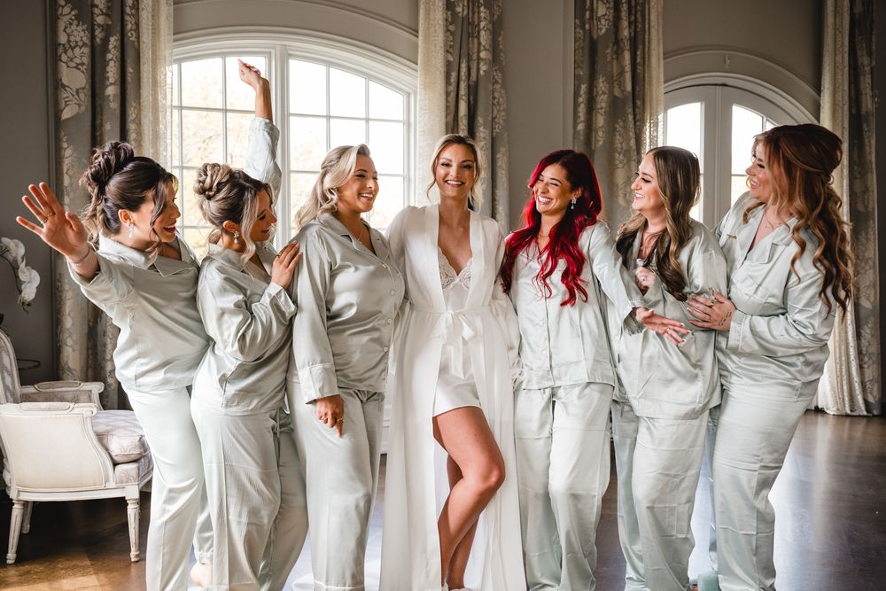 Bride in white robe poses with six bridesmaids in matching sage satin pajamas, laughing together before the wedding ceremony.