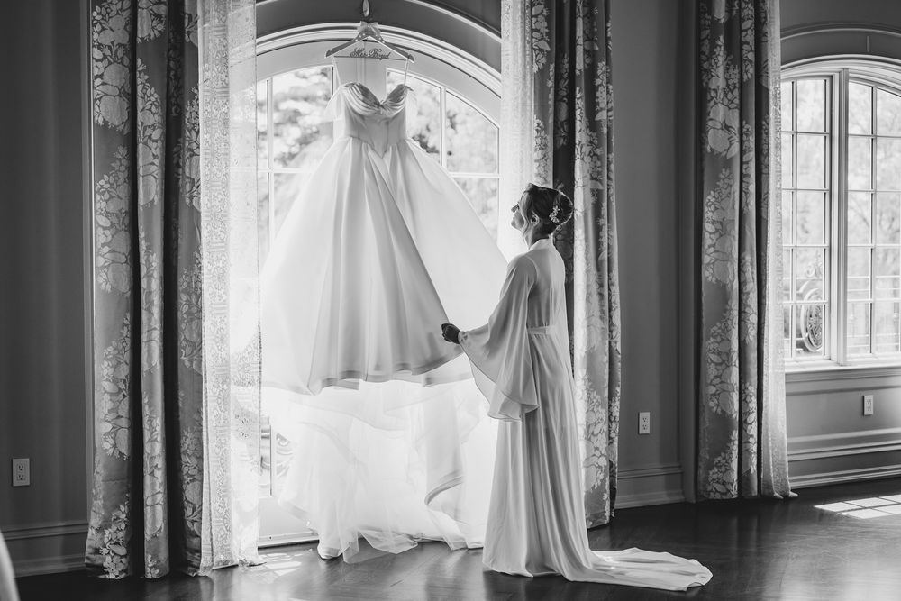 Bride in robe admiring her wedding gown hanging in an elegant window in a black and white photo.