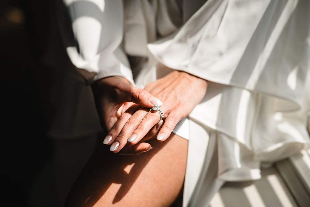 Close-up of a bride's hand wearing a diamond engagement ring, draped in soft white bridal robe fabric in natural light.