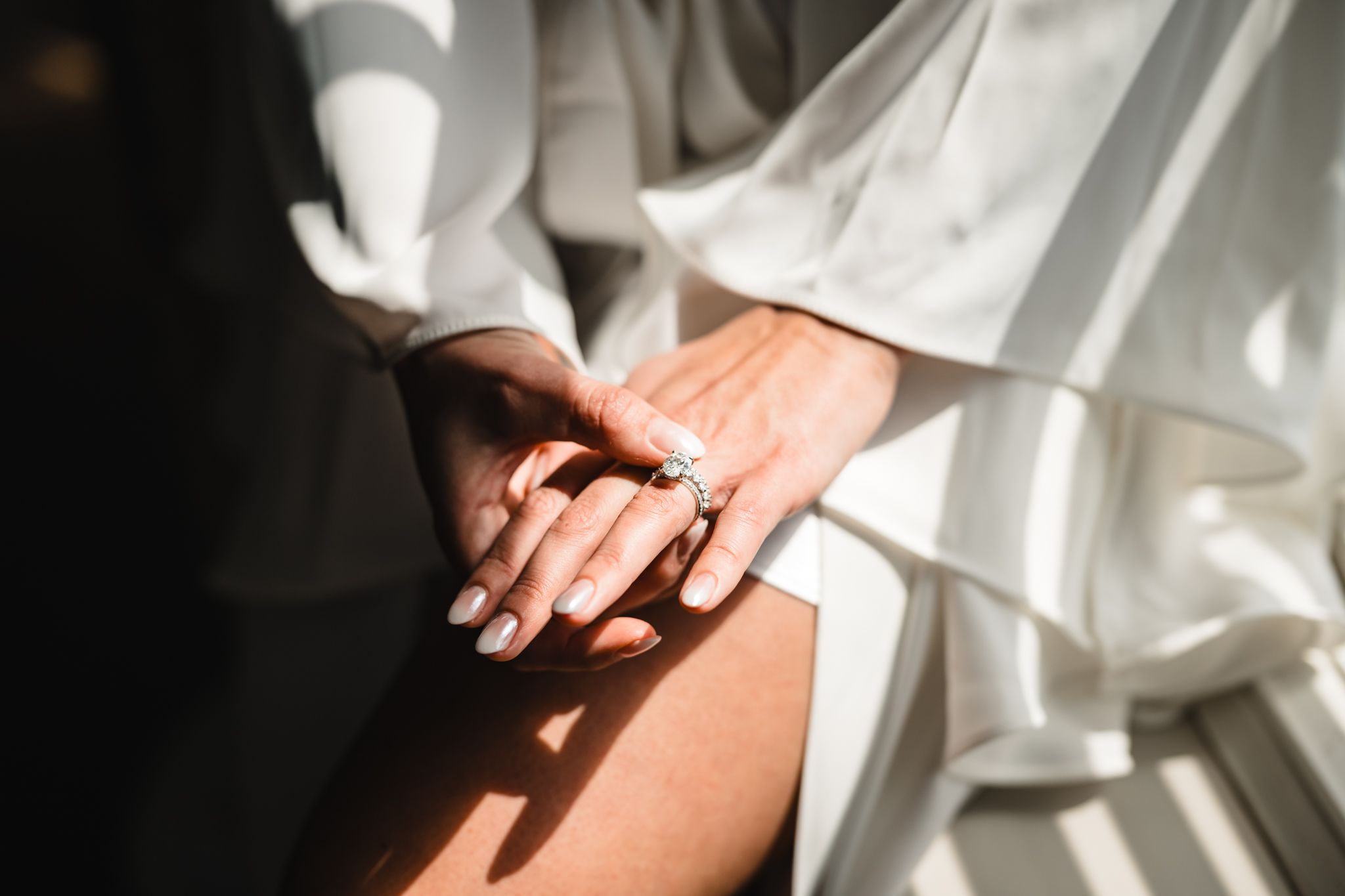 Close-up of a bride's hand wearing a diamond engagement ring, draped in soft white bridal robe fabric in natural light.