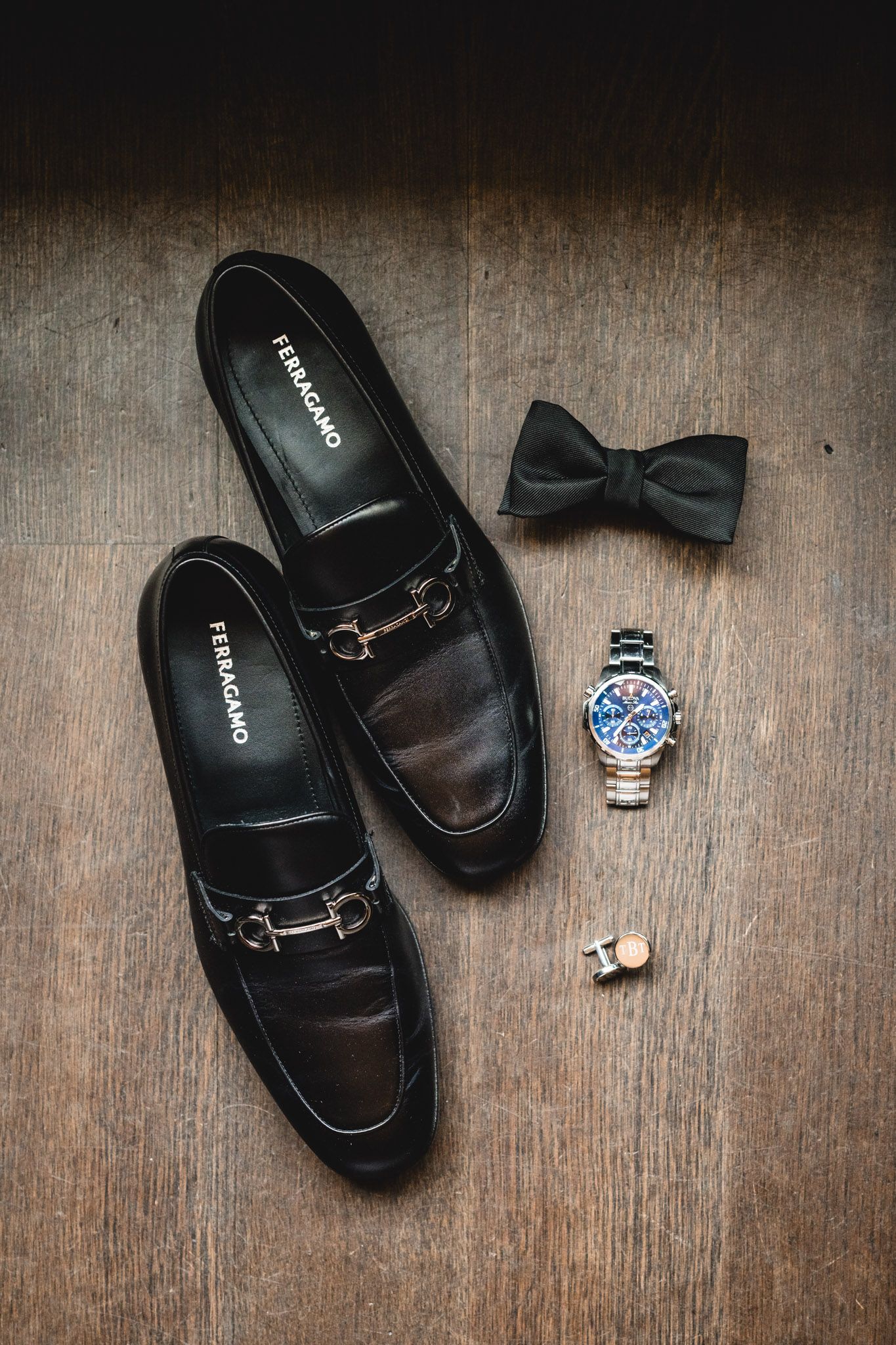 Black Ferragamo loafers displayed on wood with a bow tie, chronograph watch, and cufflinks for a groom.