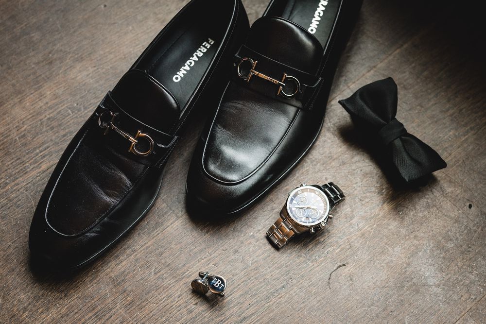 Black Salvatore Ferragamo loafers beside a gold watch, cufflinks, and black bow tie on wooden floor.