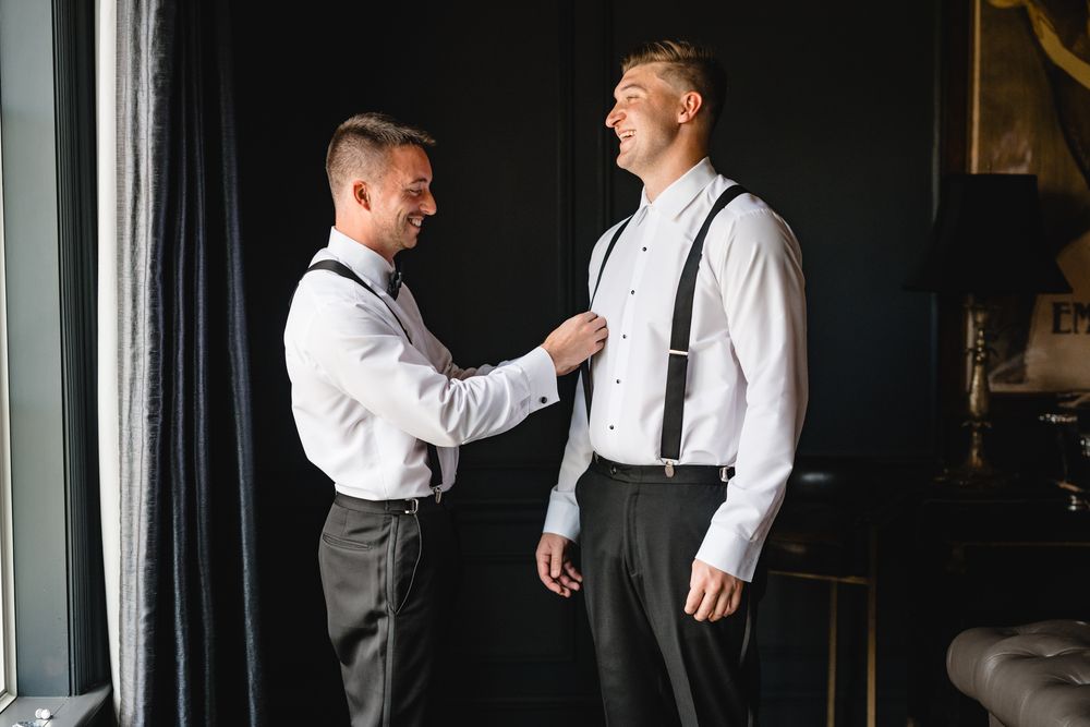Two groomsmen in white shirts and black suspenders help each other prepare for a wedding ceremony.