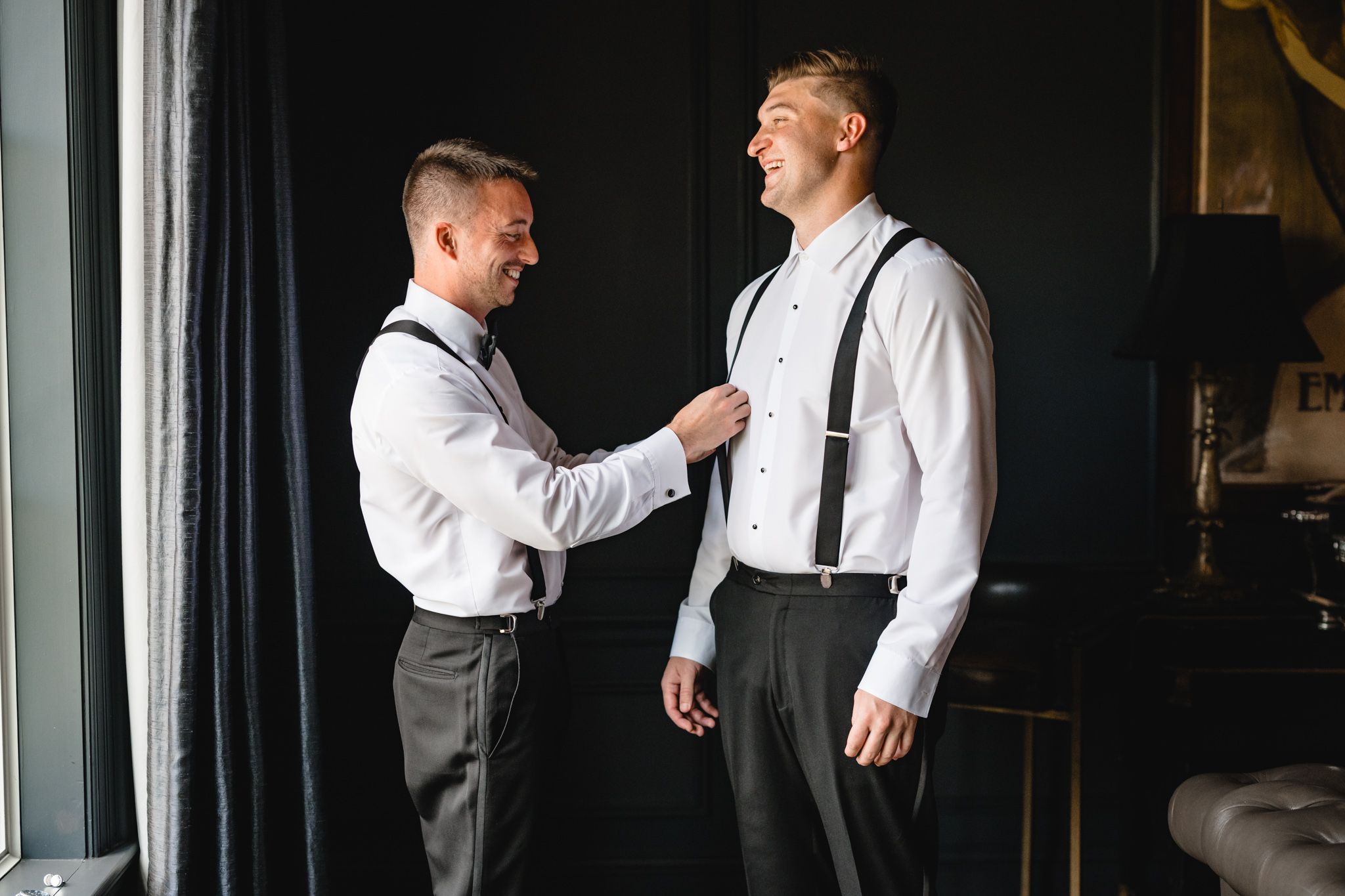 Two groomsmen in white shirts and black suspenders help each other prepare for a wedding ceremony.