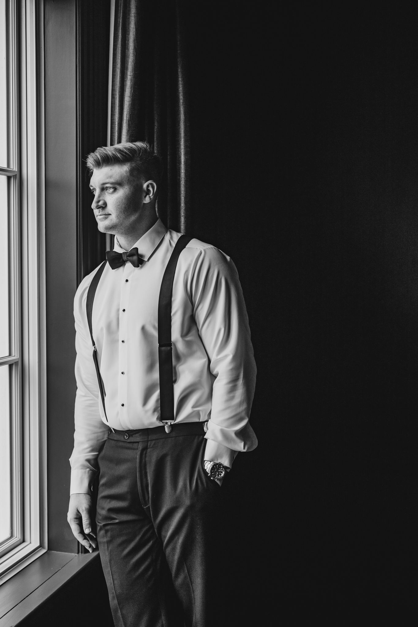 Young groom in white dress shirt with suspenders and bow tie standing by window in black and white portrait.