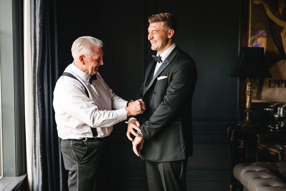 An older man helps a young groom in a black tuxedo adjust his jacket before the wedding ceremony.