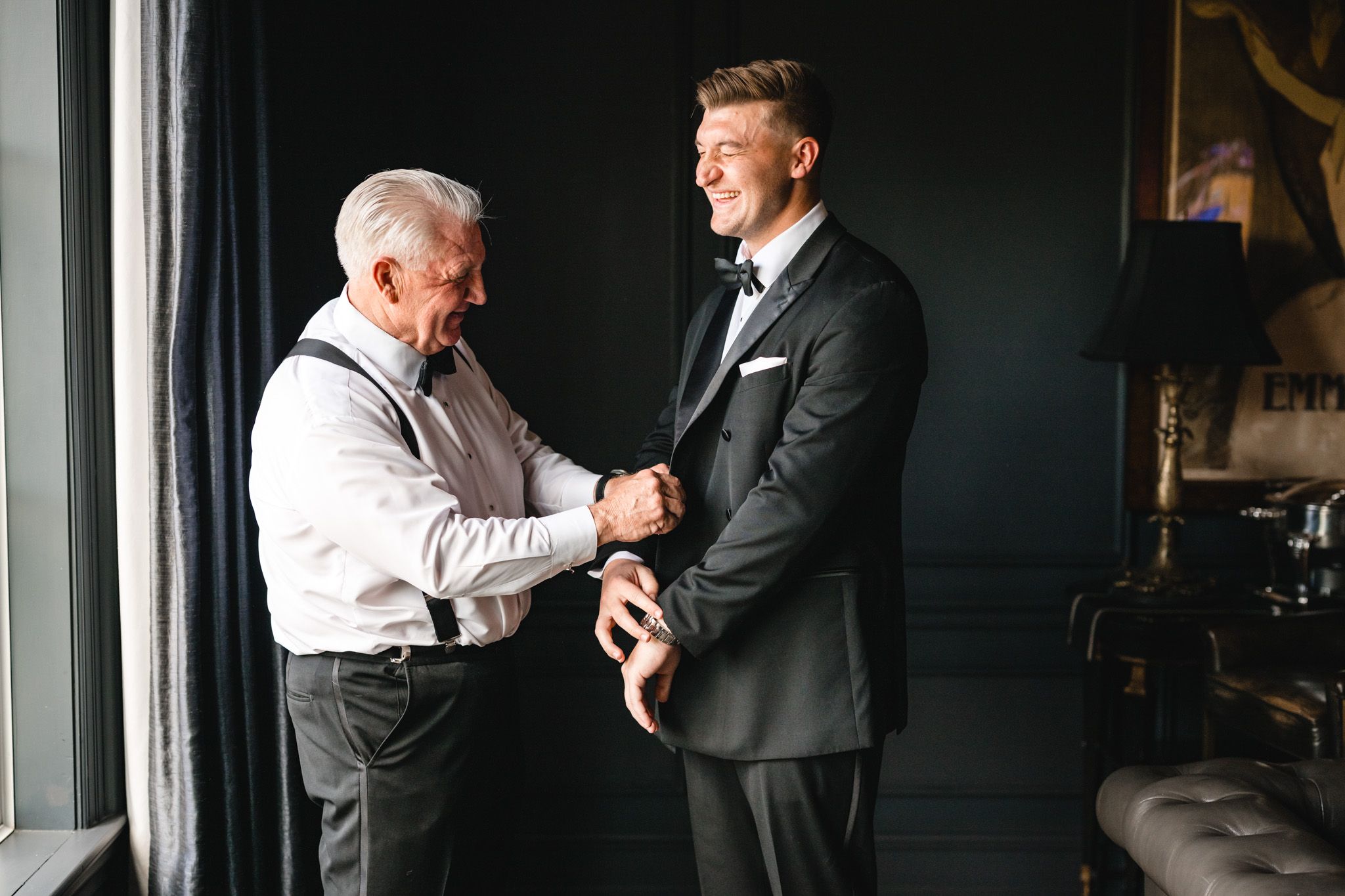 An older man helps a young groom in a black tuxedo adjust his jacket before the wedding ceremony.