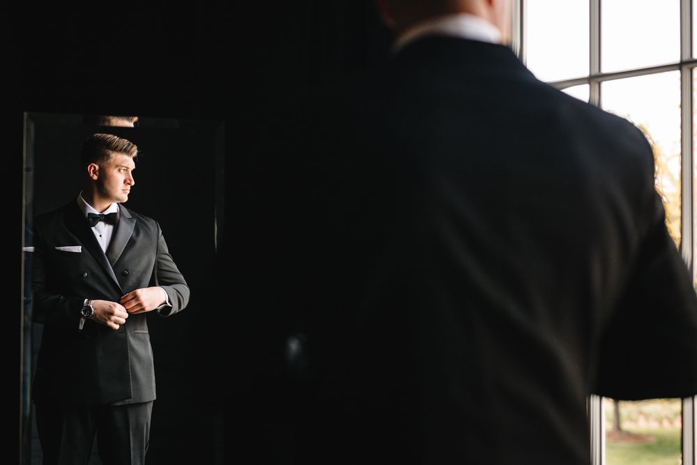Elegant groom in black tuxedo adjusting jacket while viewing reflection in mirror before wedding.