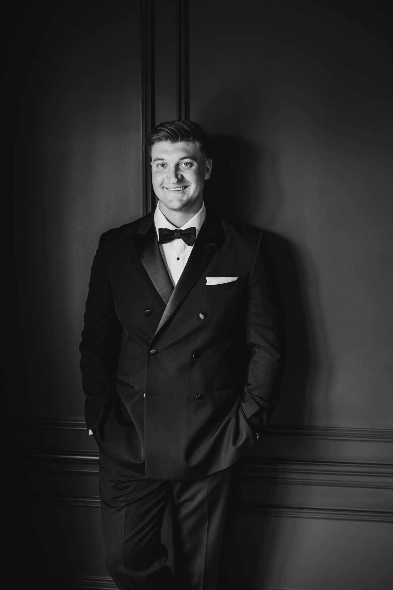 Handsome groom smiling in double-breasted black tuxedo with bow tie in dramatic black and white portrait.