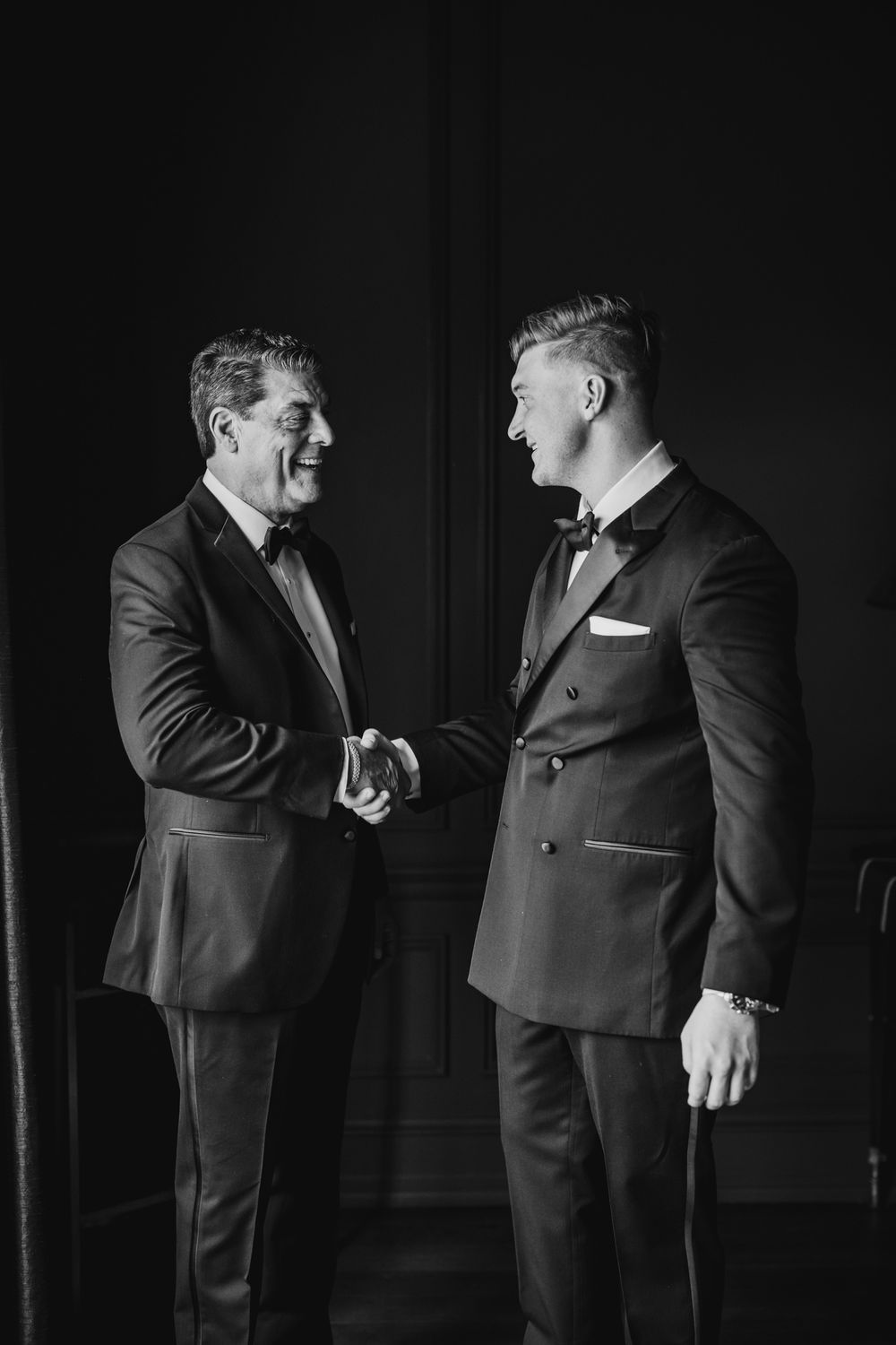 Two men in formal black tuxedos sharing a warm handshake in elegant black and white wedding photo.