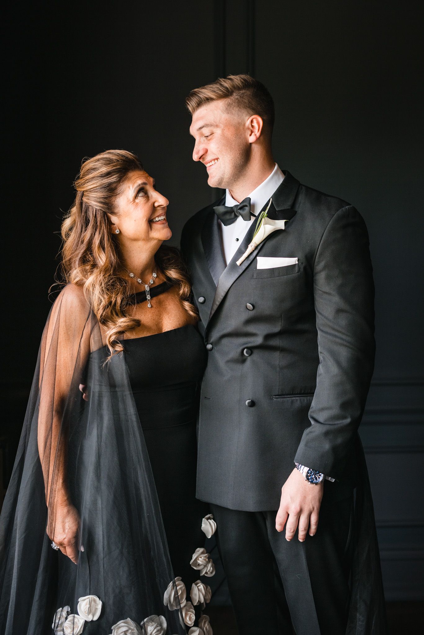 Elegant couple in matching black formal attire smiling at each other against a dark background.
