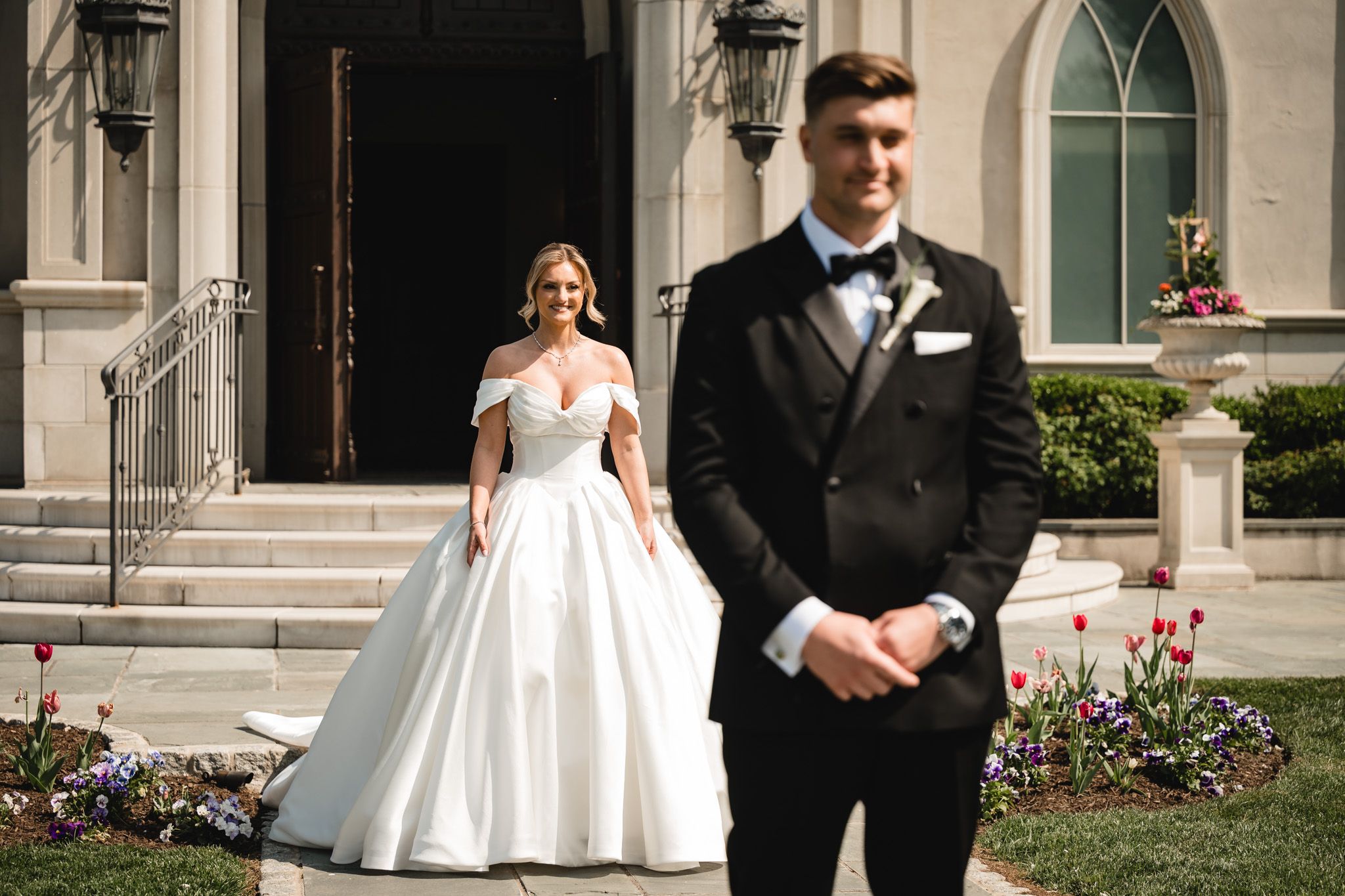 Bride in off-shoulder ball gown stands behind groom in tuxedo outside elegant stone church.