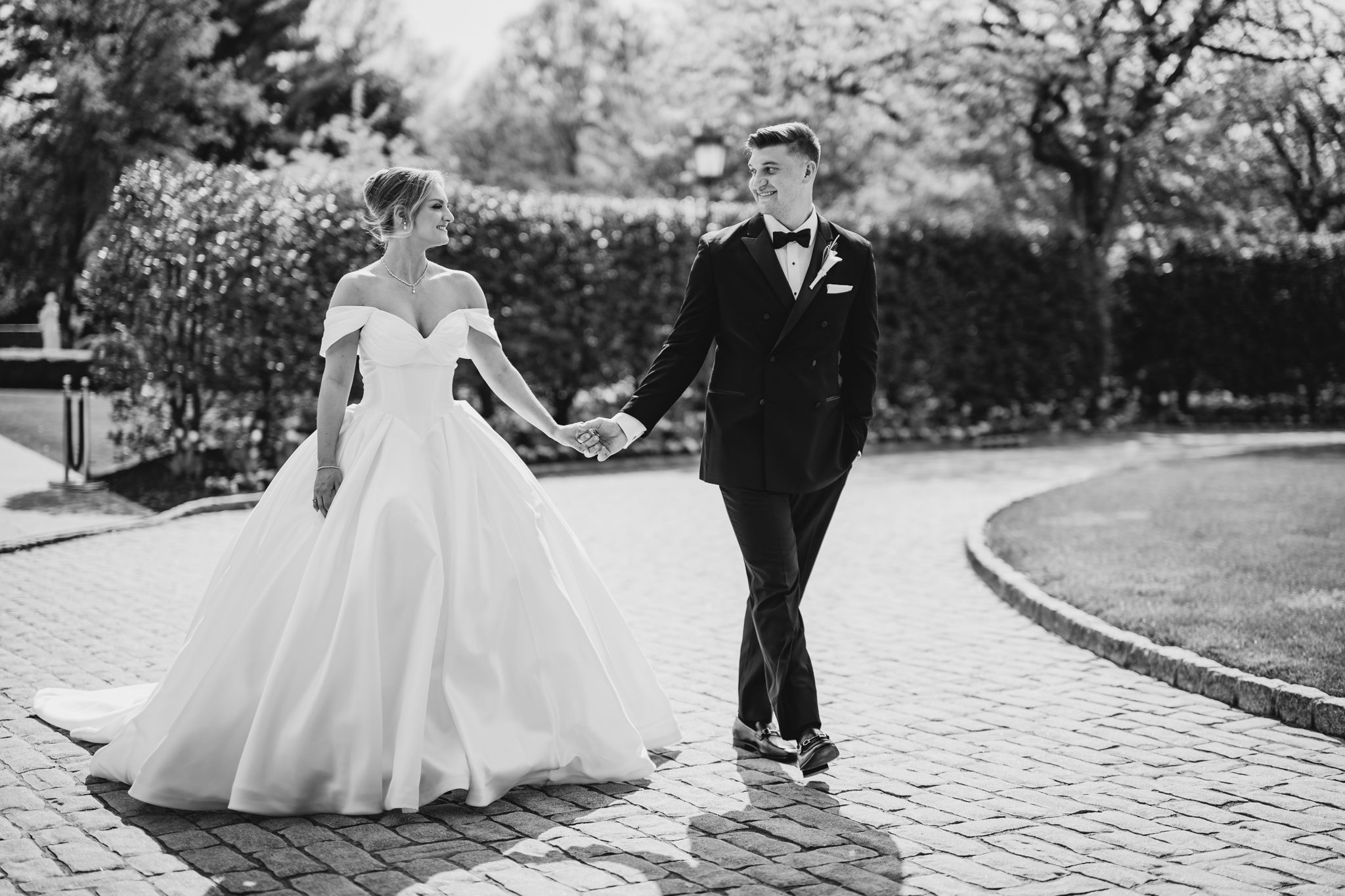 Black and white photo of bride in ball gown and groom holding hands on brick pathway.