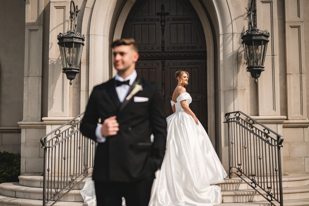 A bride in a white ball gown and groom in black tuxedo pose on church steps for wedding portraits.