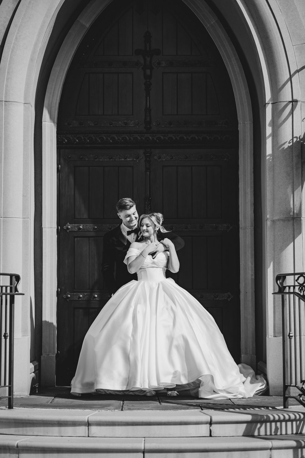 Bride and groom embrace on church steps in elegant black and white wedding portrait.