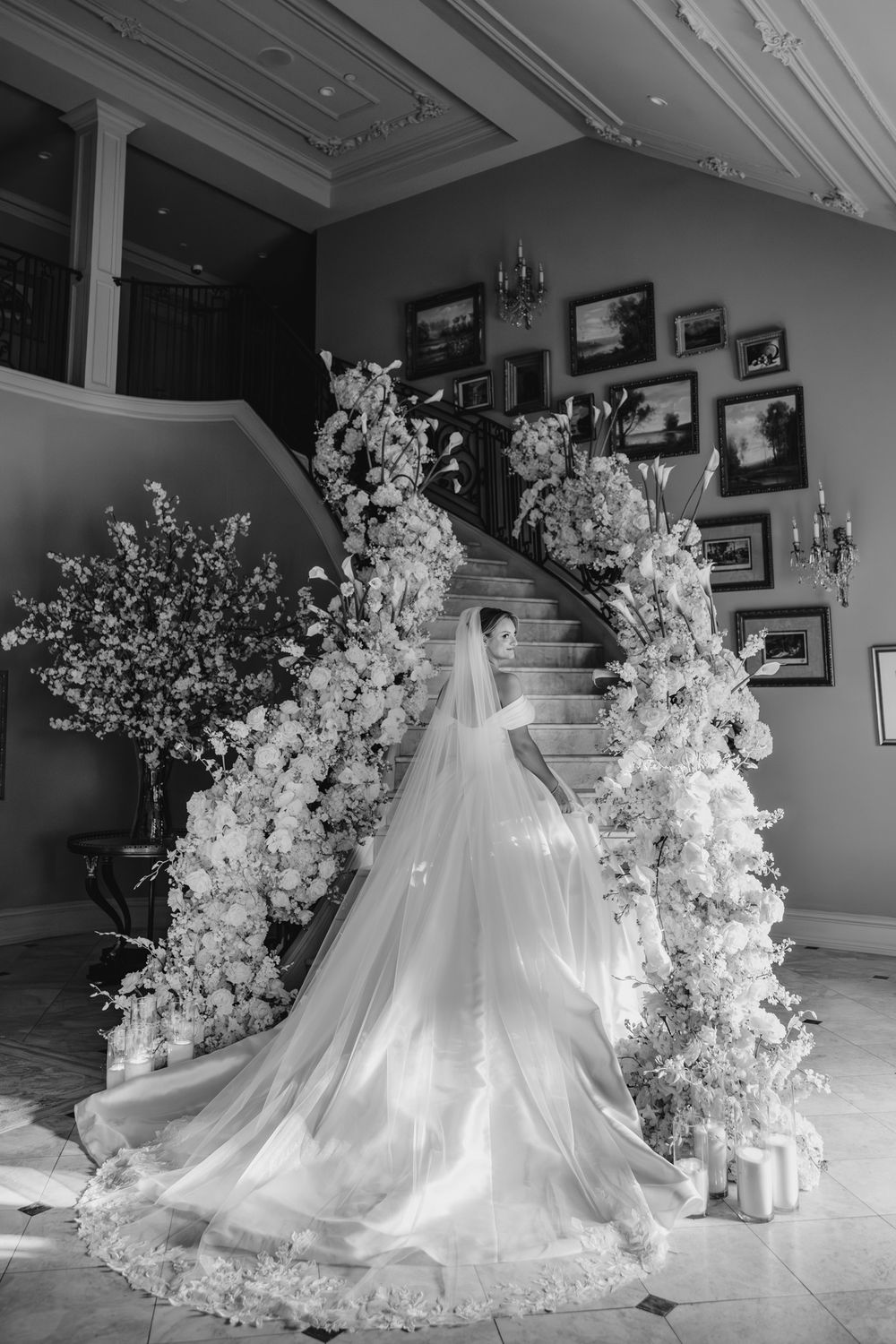 Bride in flowing gown and veil poses on floral-adorned staircase in black and white.