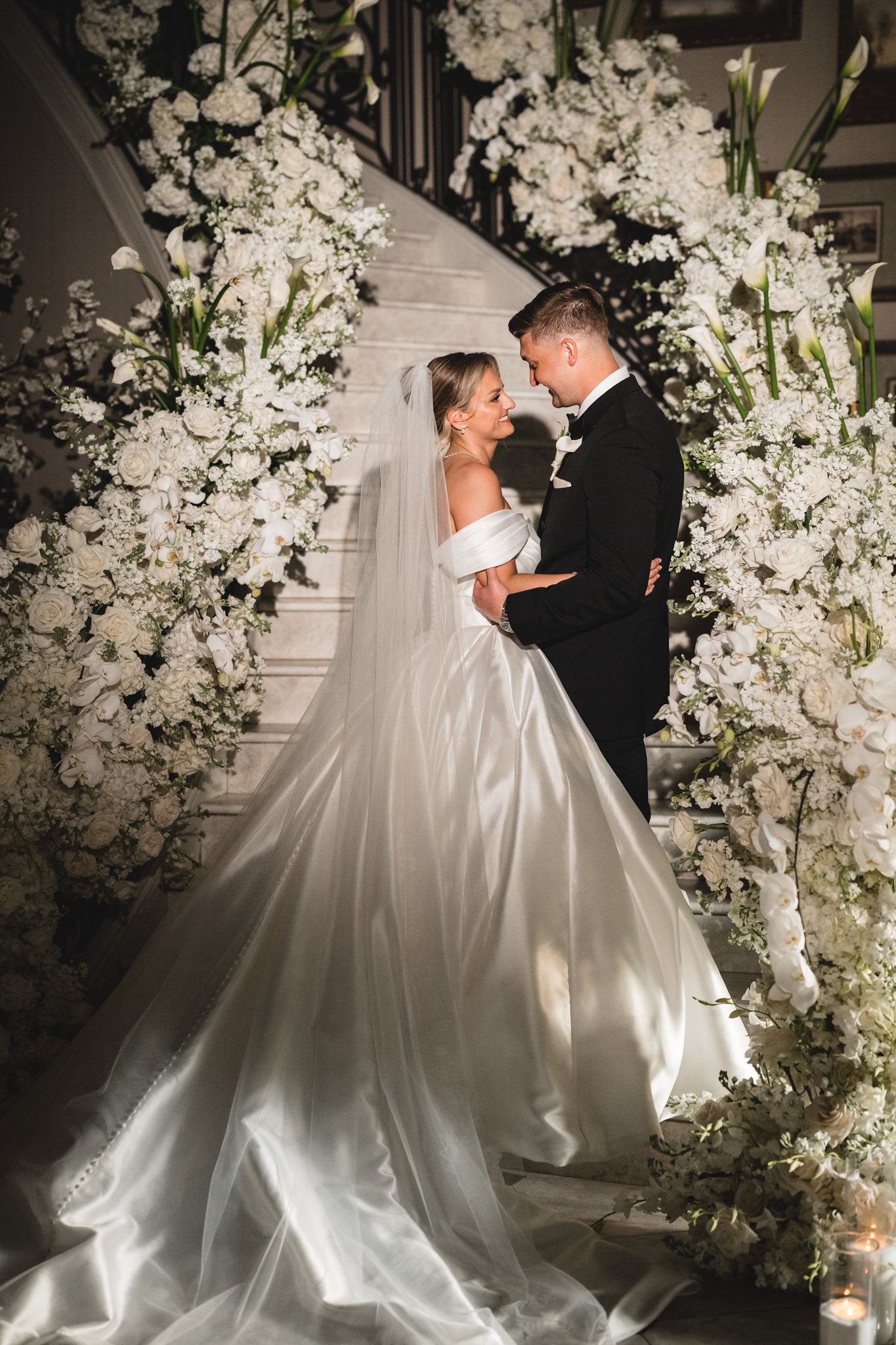 Bride in a flowing satin gown and groom stand surrounded by lush white floral arrangements.