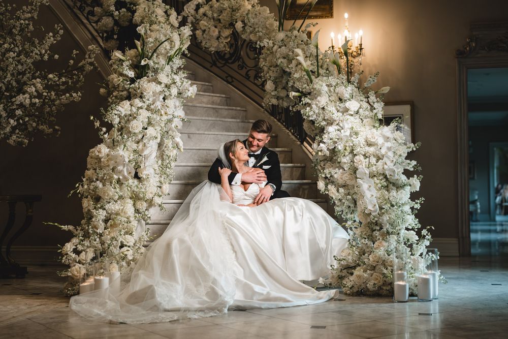 Bride and groom share romantic moment seated beneath lush white floral arch on grand staircase.