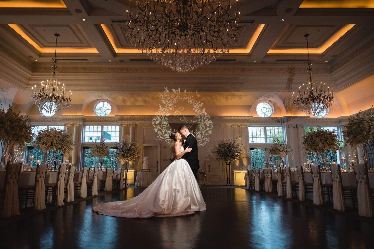 Bride and groom share romantic kiss under grand chandeliers in elegant ballroom.