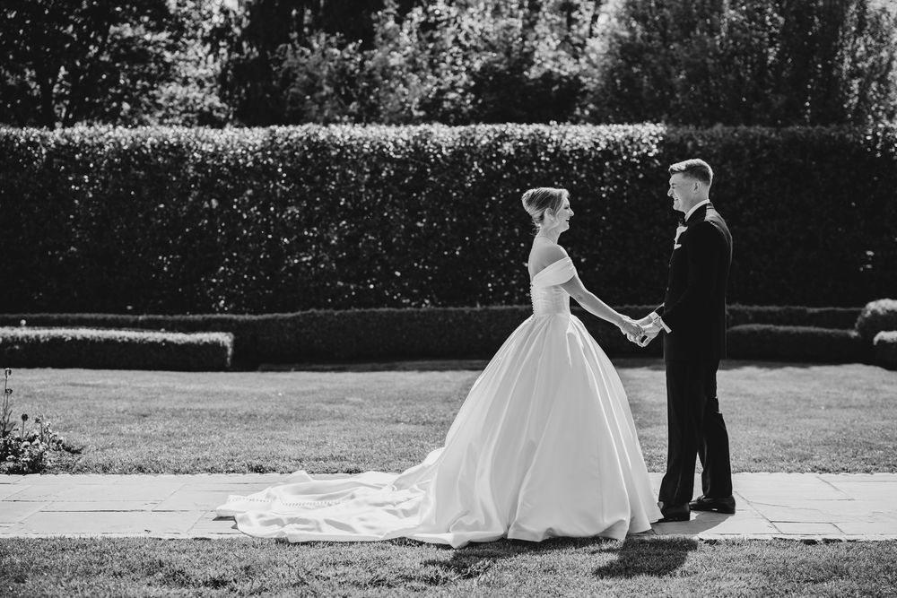 Bride and groom hold hands in black and white photo on manicured garden path.