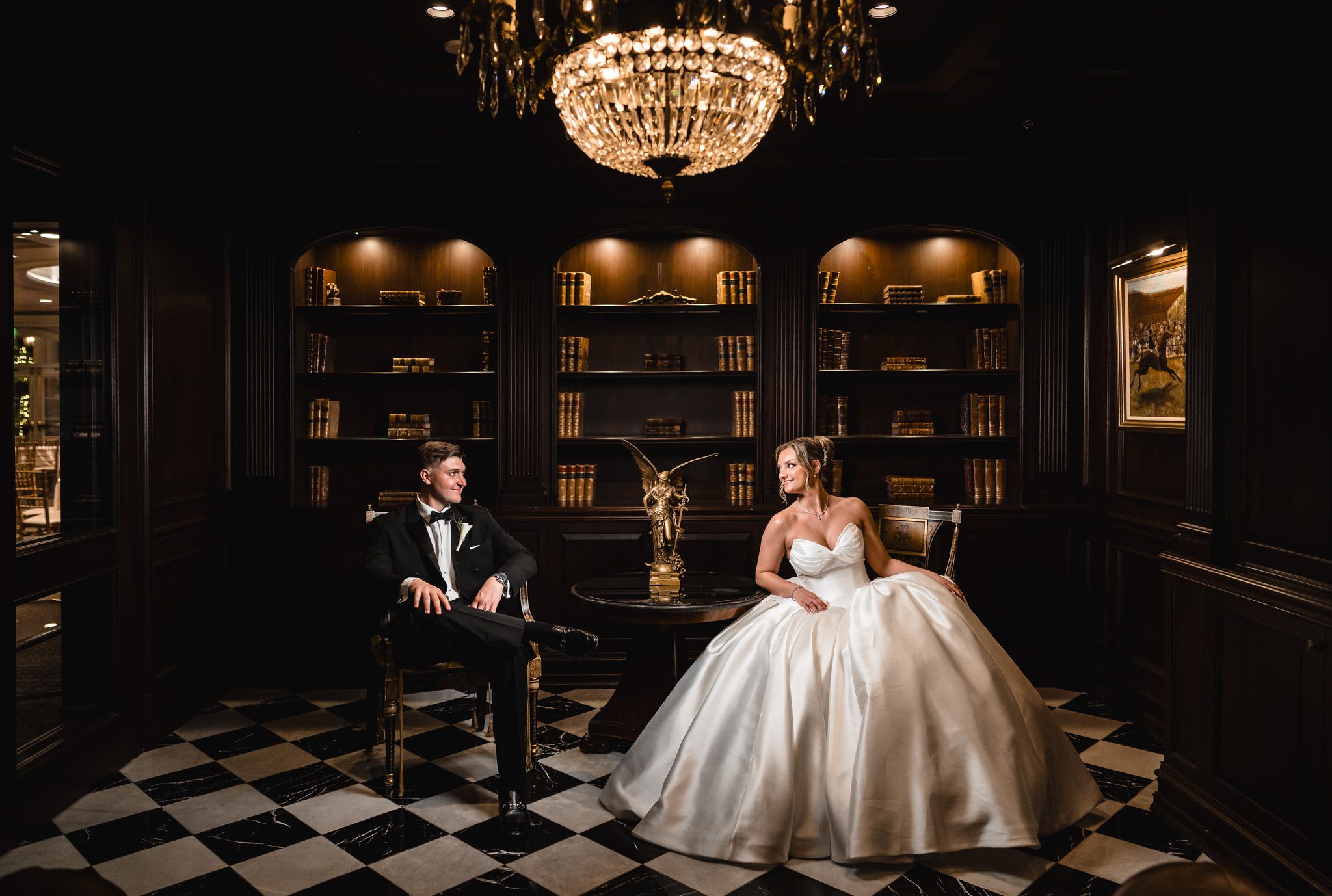 Bride and groom seated in a dramatic dark library room with chandelier and checkered floor.