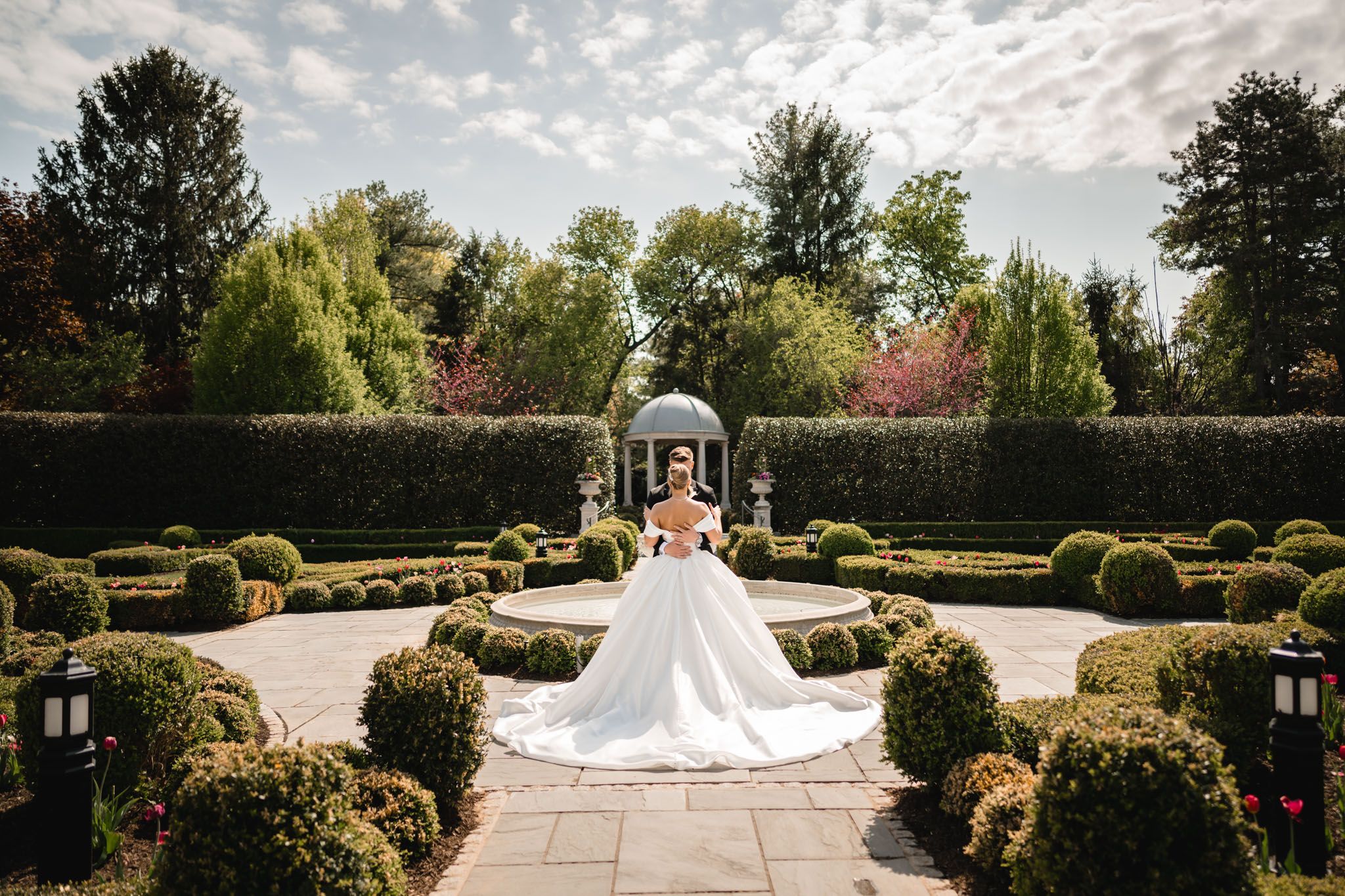 Bride in a white ball gown poses in a formal garden with manicured hedges and a gazebo backdrop.