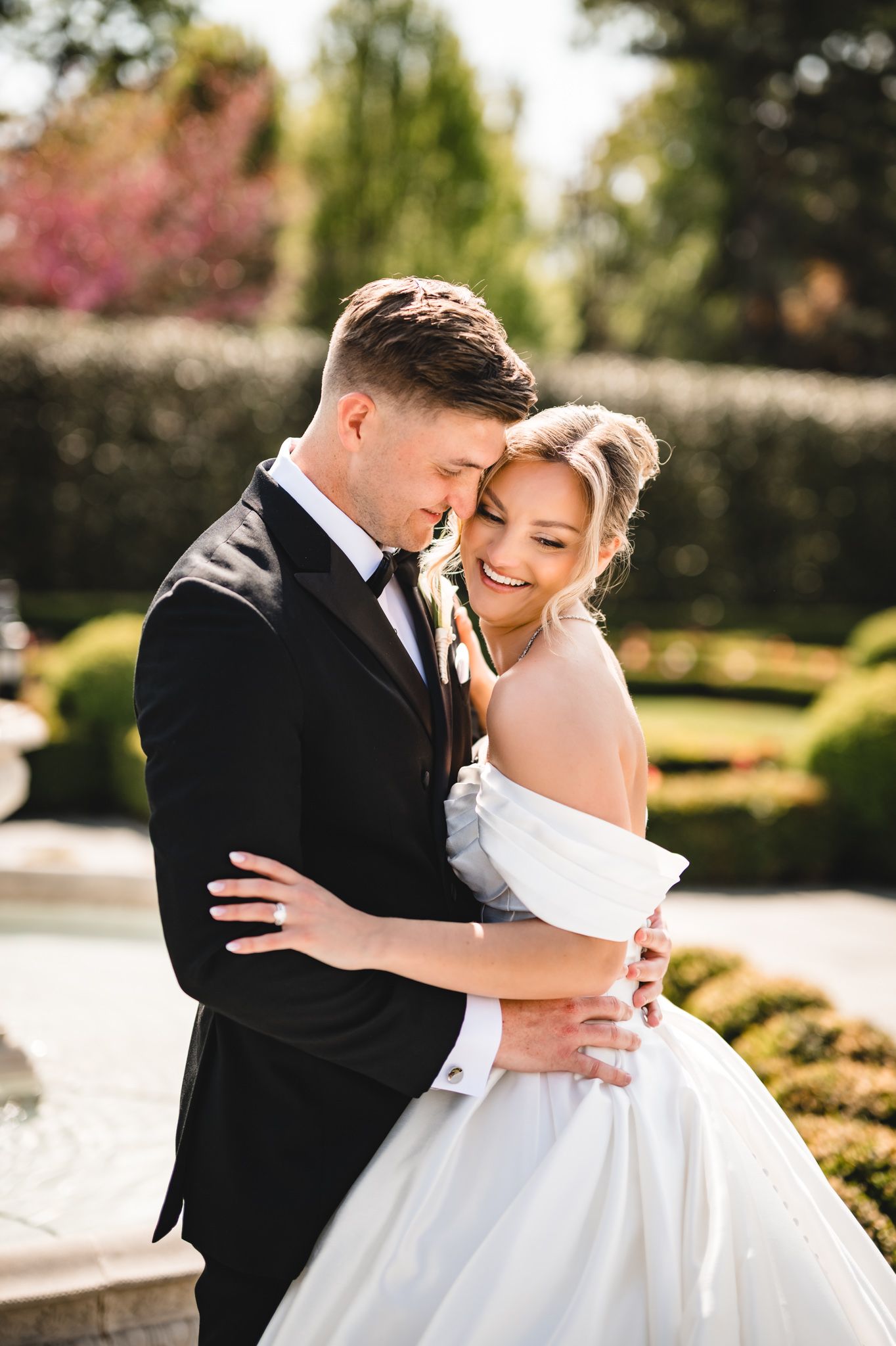Joyful bride and groom embracing in sunlit garden with bride wearing off-shoulder white ball gown.