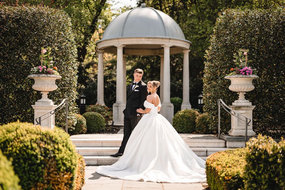 Bride and groom posing elegantly in a lush garden with a classic gazebo backdrop.