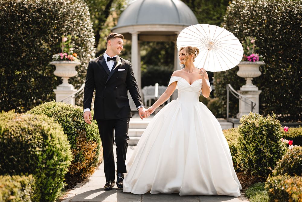 Bride and groom walking through formal garden with bride holding white parasol and wearing ball gown.