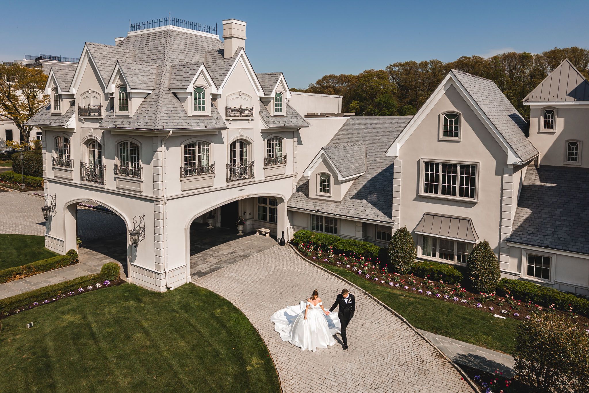 Aerial view of bride and groom walking toward elegant white mansion wedding venue with manicured grounds.