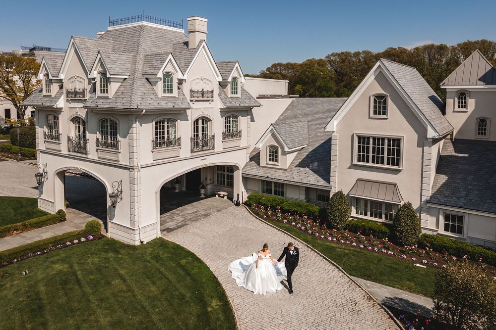 Aerial view of bride and groom walking toward elegant white mansion wedding venue with manicured grounds.