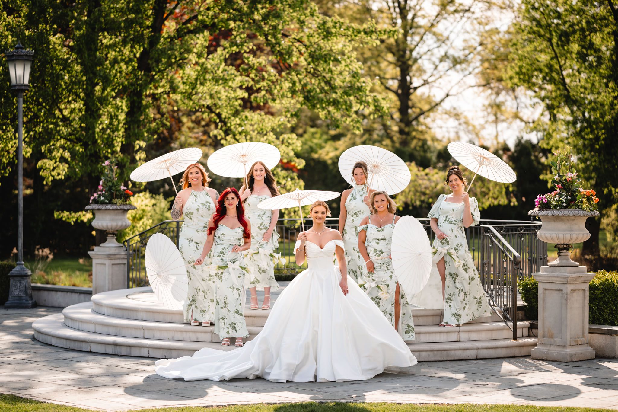 A bride in white gown poses with bridesmaids in floral dresses holding parasols on garden steps.