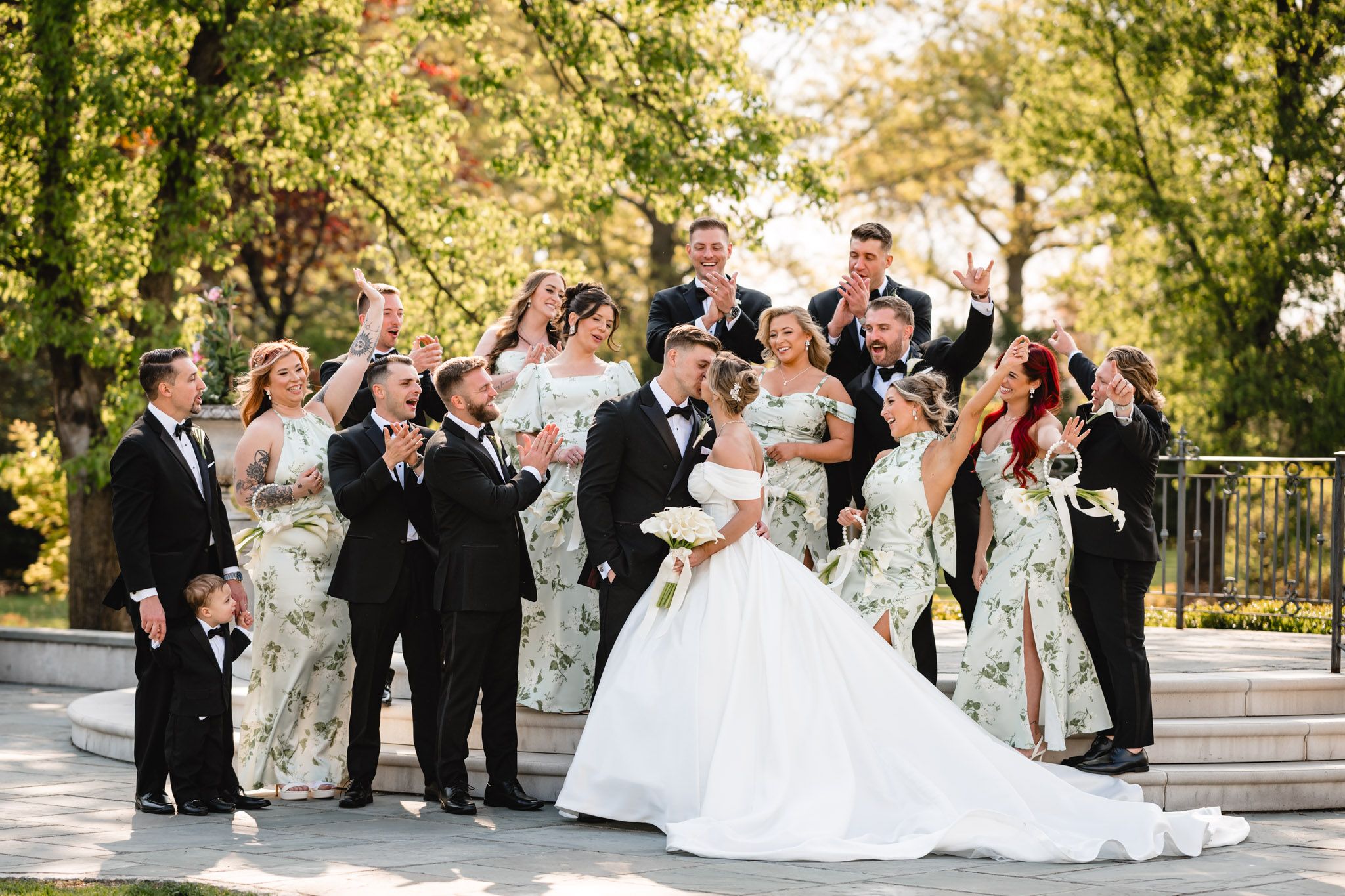 Lively wedding party celebrating outdoors with bridesmaids in floral dresses and groomsmen in black tuxedos.