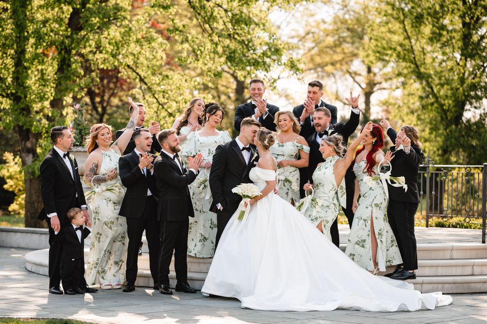 Lively wedding party celebrating outdoors with bridesmaids in floral dresses and groomsmen in black tuxedos.